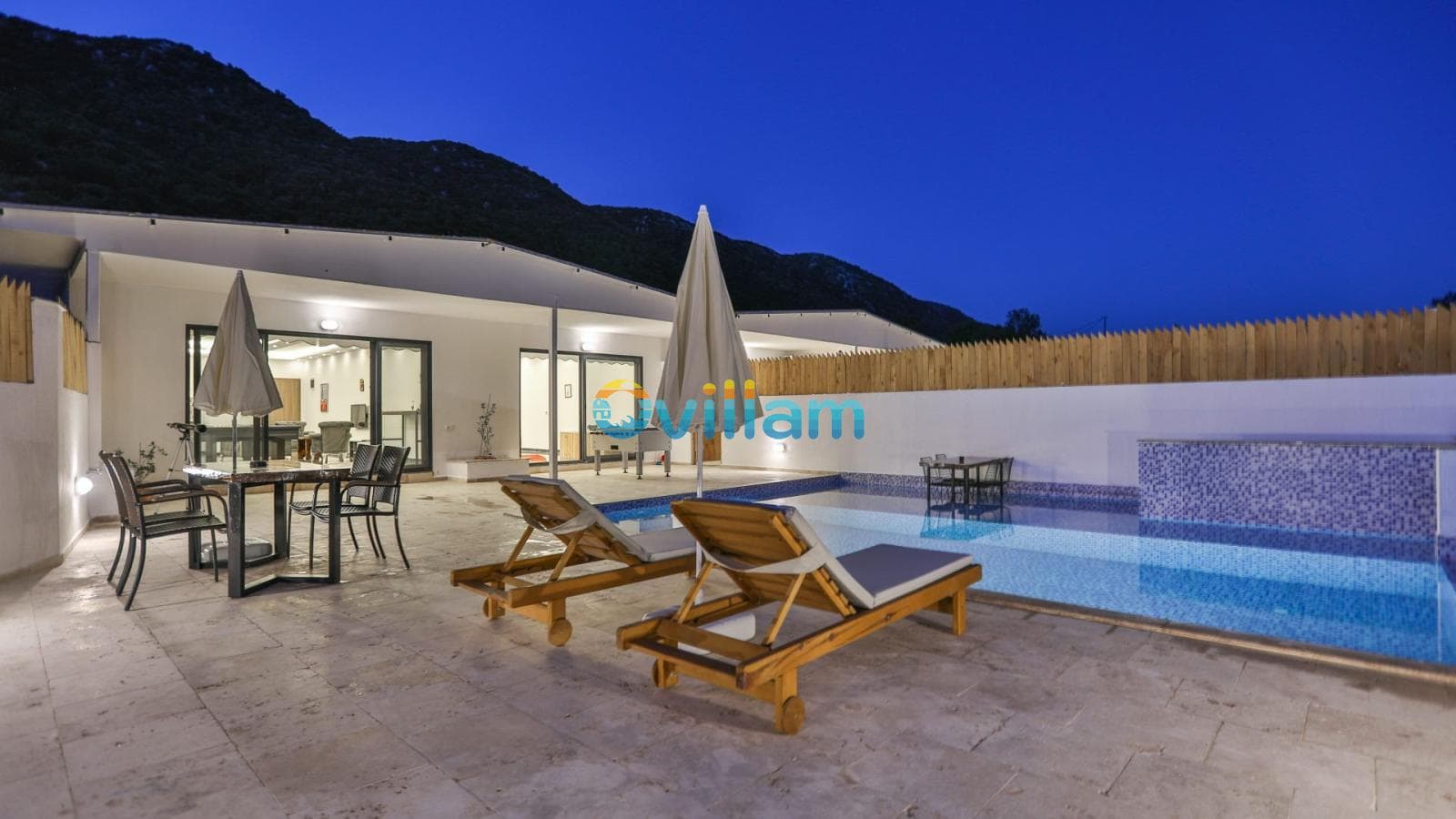 Gece aydınlatmalı havuz kenarında şezlonglar, masa ve sandalyeler bulunan taş zeminli villa terası.