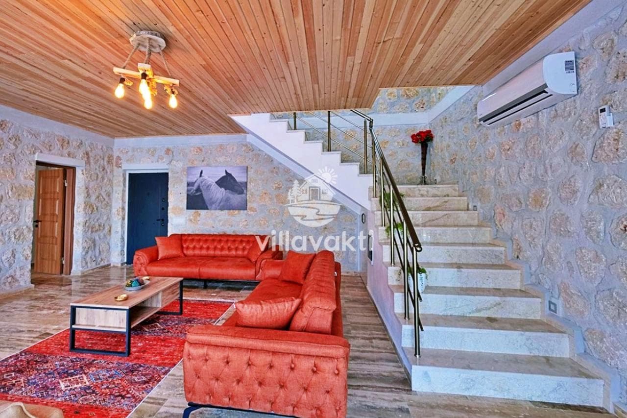 Ahşap tavanlı oturma odasında kırmızı koltuklar, desenli halı ve üst kata çıkan mermer merdivenler var.