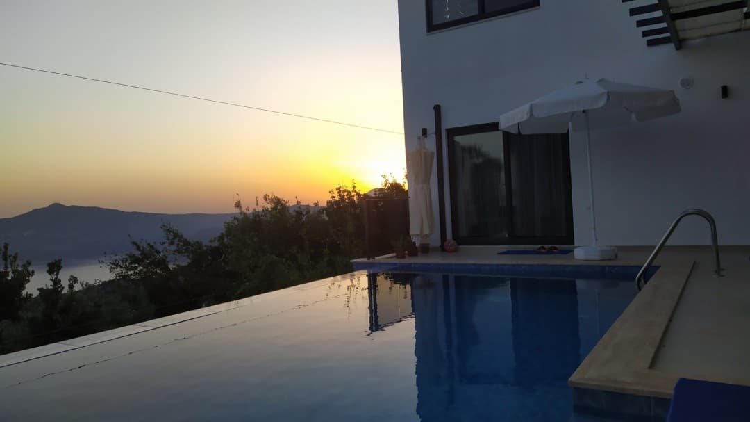 Gün batımında sonsuzluk havuzu ve yanında beyaz şemsiyesi bulunan beyaz dış cepheli villa.