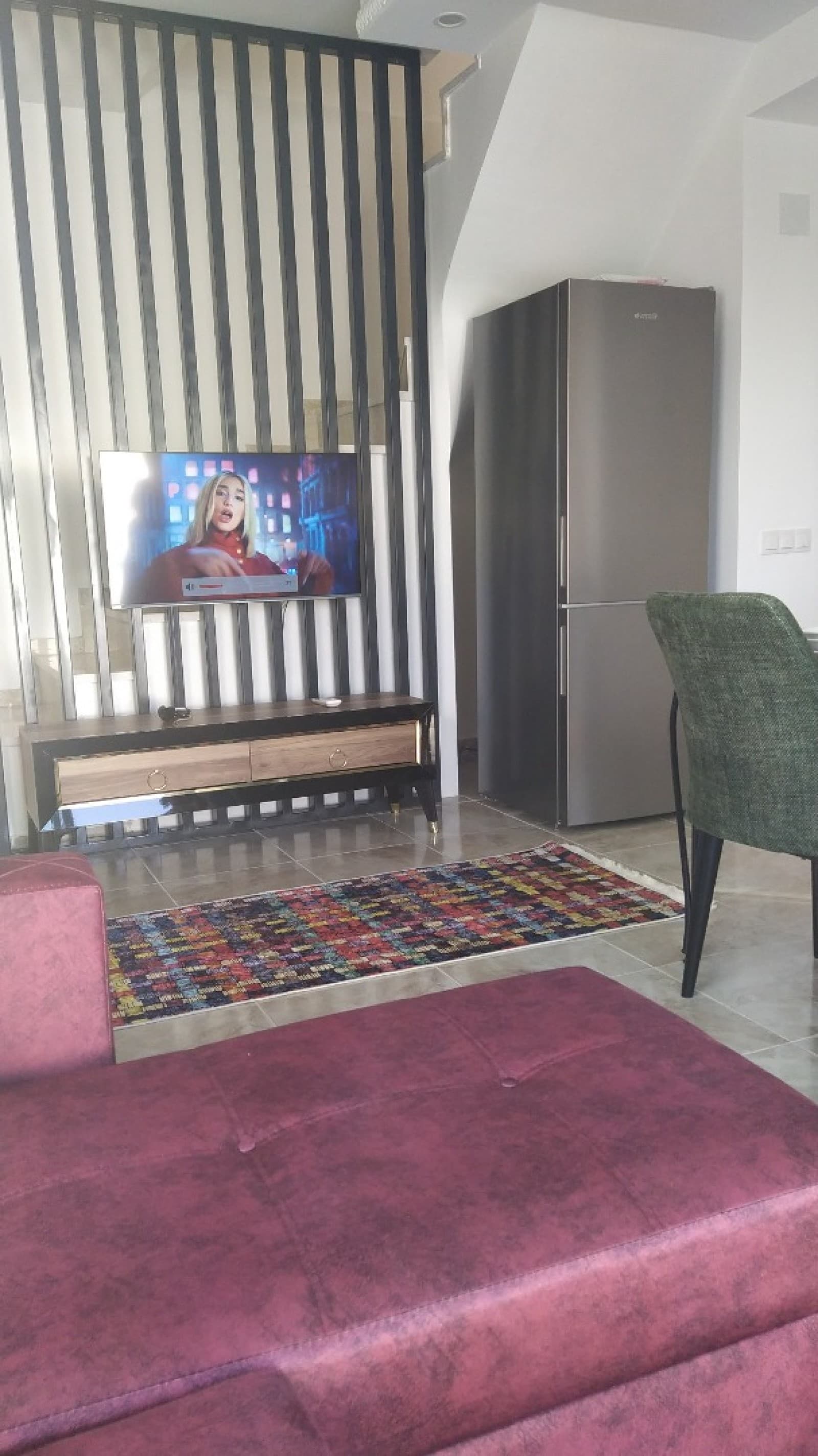 Dikey çıtalı duvar panelinde asılı televizyon ve önünde bordo koltuk.