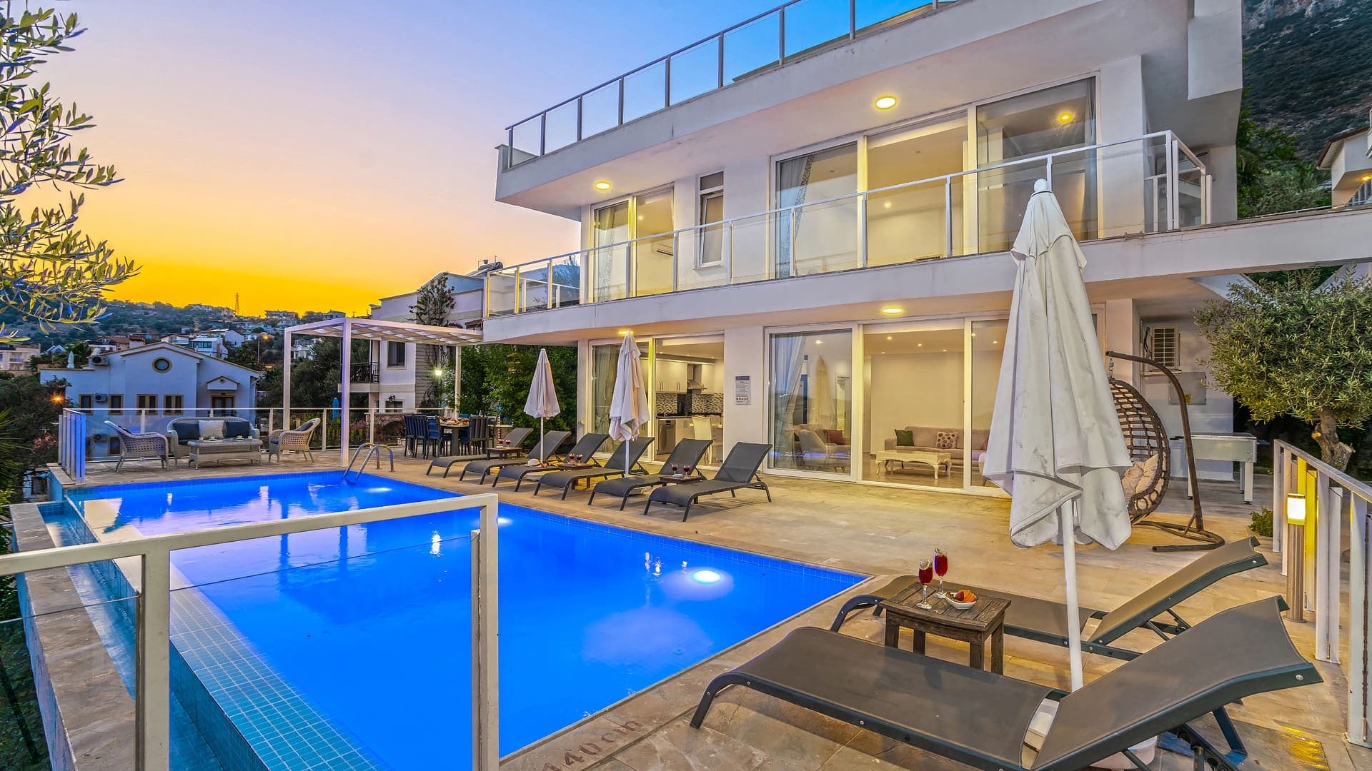 Gün batımı manzaralı, havuz başında şezlonglar ve şemsiyeler bulunan iki katlı villa dış cephesi.