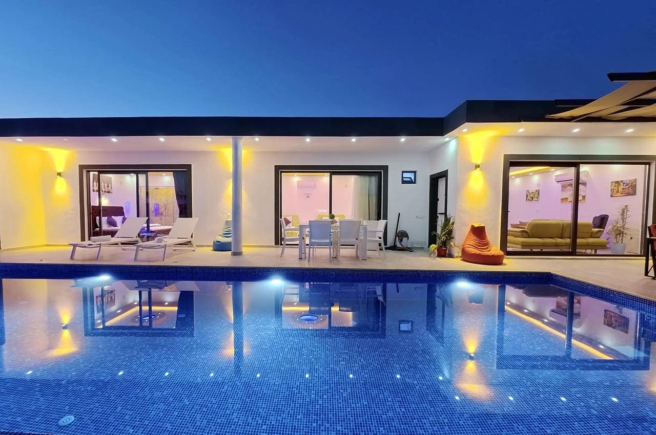 Gece aydınlatmalı havuz ve önünde oturma grubu bulunan modern villa dış cephesi.