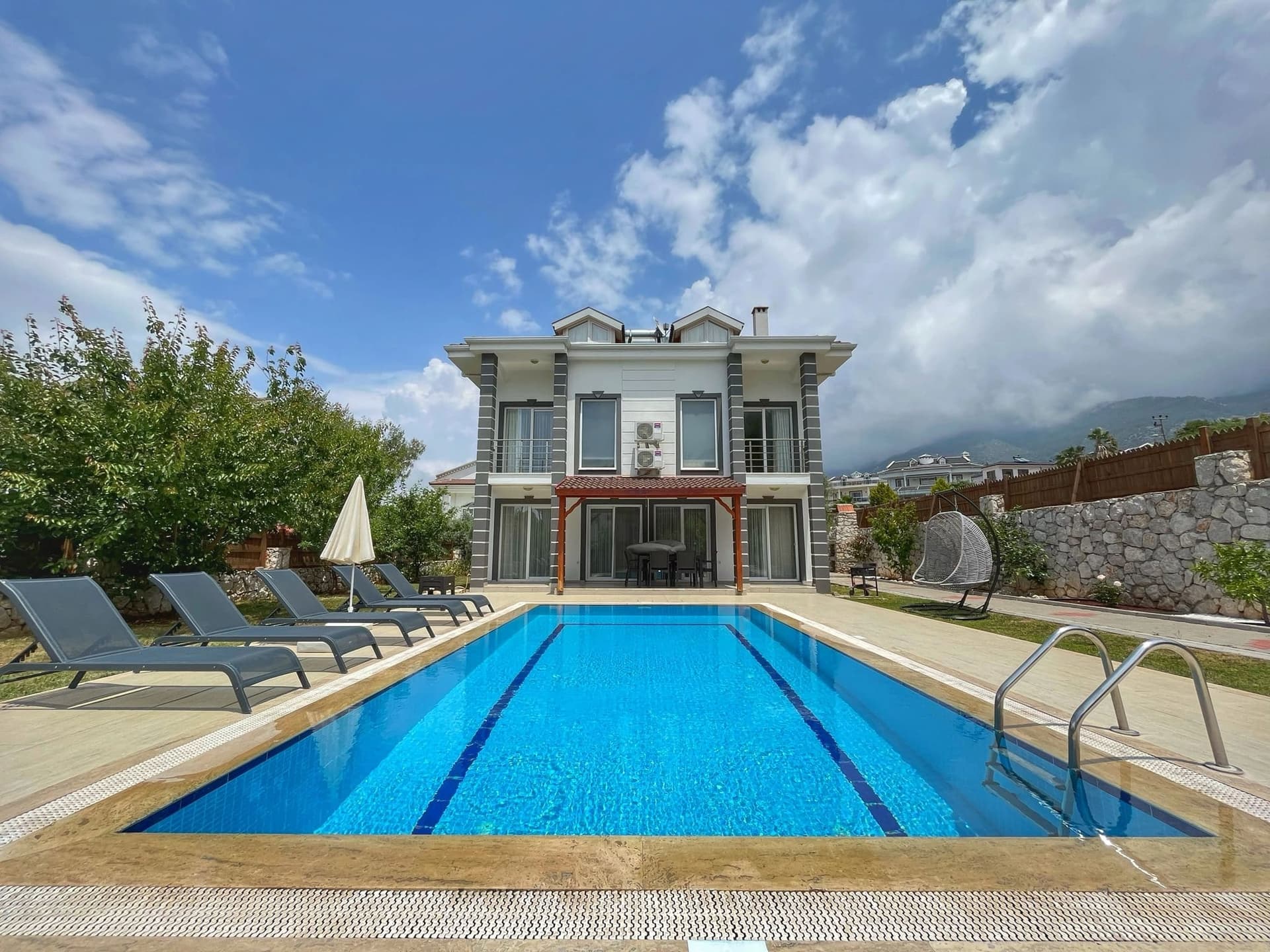 Önünde şezlonglar ve şemsiye bulunan, mavi sularla dolu dikdörtgen bir yüzme havuzu ve arkasında villa.