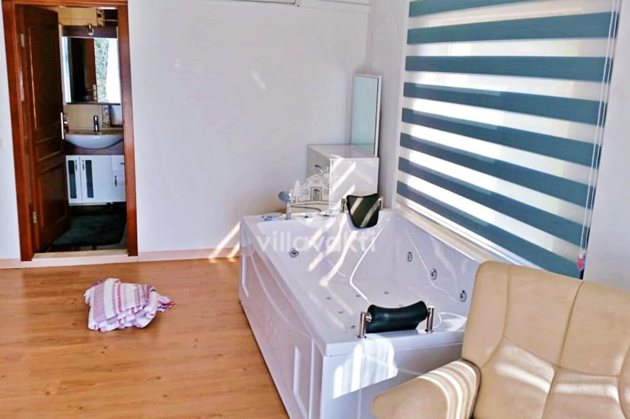 Ahşap zeminli iç mekânda çift kişilik jakuzi, berjer ve arkada görünen banyo kapısı.