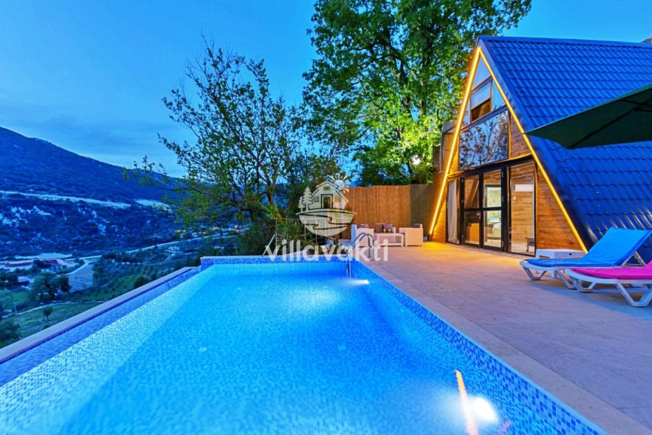 Akşam saatlerinde aydınlatılmış A-tipi ahşap villa, havuz kenarında şezlonglar ve dağ manzarası.