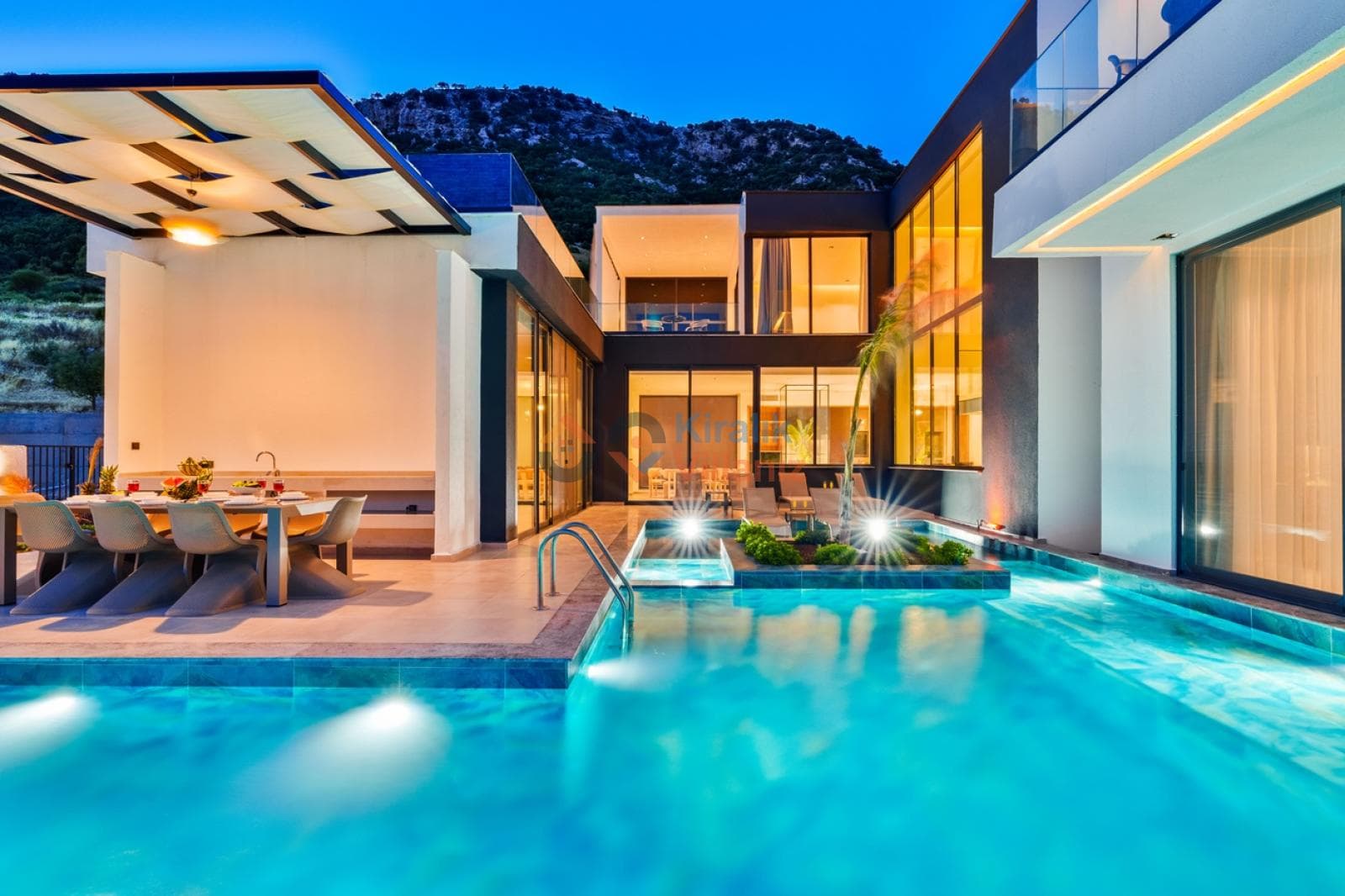 Akşam ışıklandırmalı modern villa, havuz kenarında yemek masası ve cam cepheli mimari.