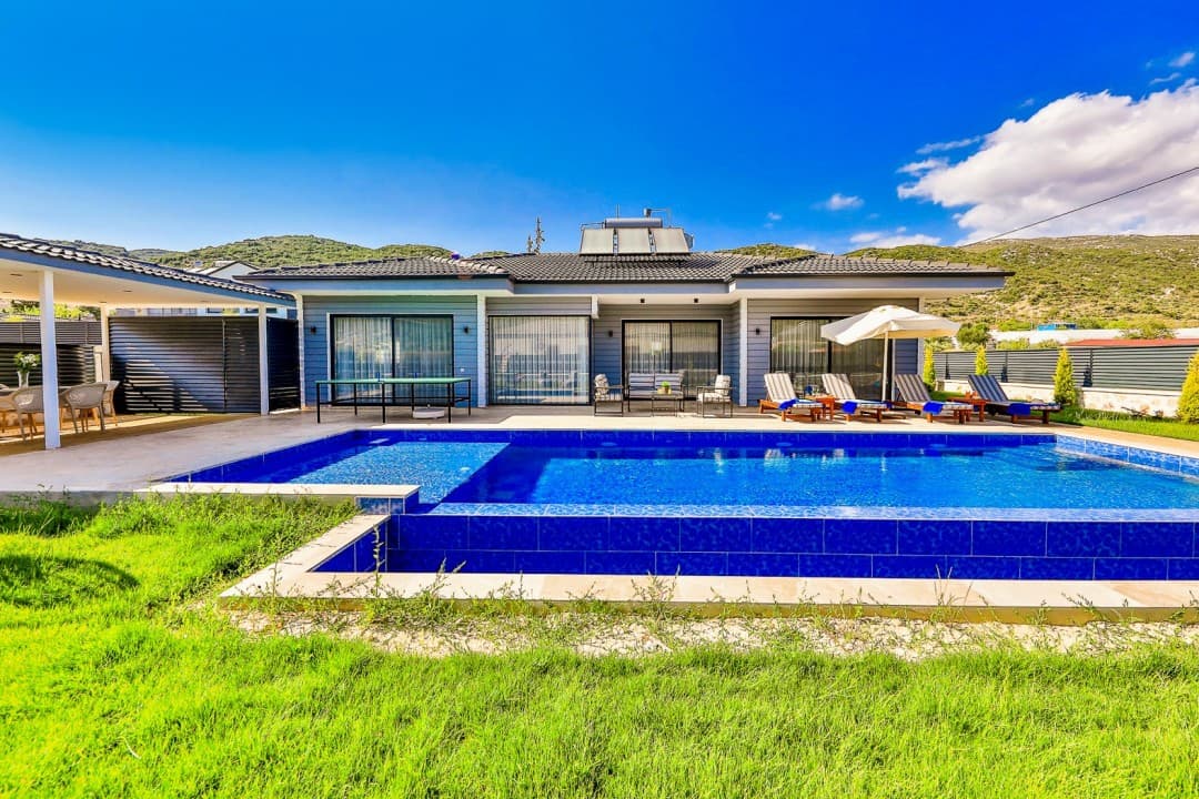 Önünde yüzme havuzu, şezlonglar ve masa tenisi bulunan, bahçeli tek katlı modern villa.