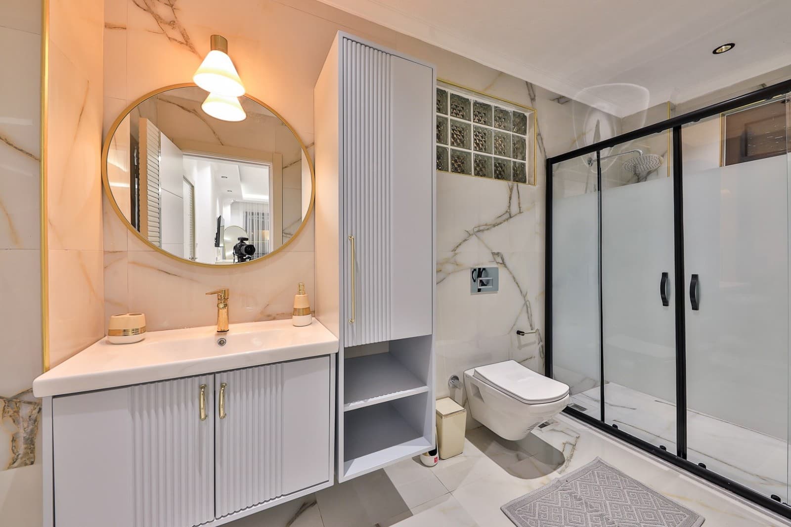 Mermer kaplı banyoda altın çerçeveli yuvarlak ayna, beyaz lavabo dolabı ve cam duş kabini yer alıyor.