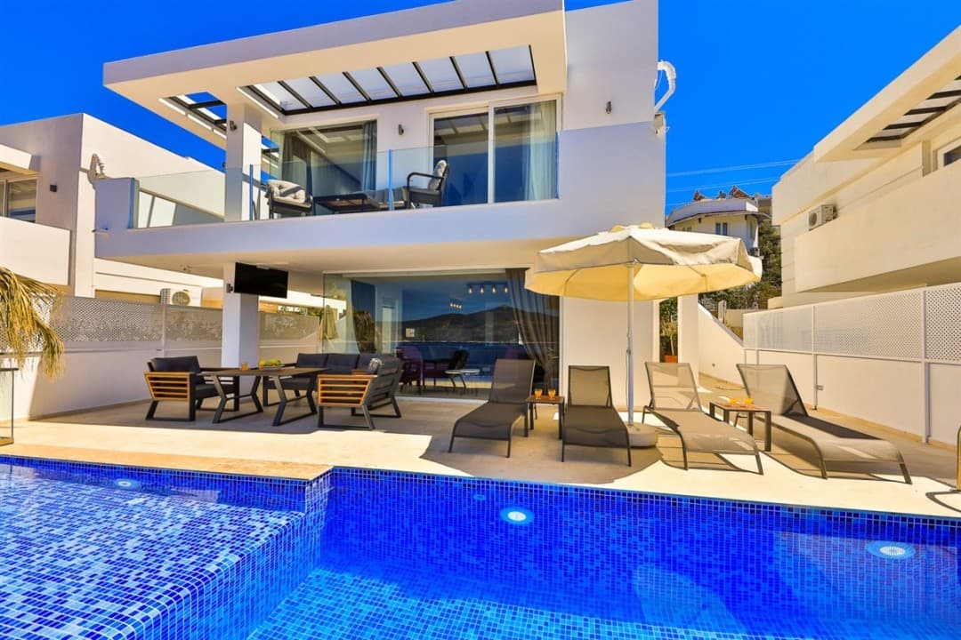 Önünde mavi mozaik kaplı havuz, şezlonglar ve oturma grubu bulunan iki katlı modern beyaz villa.