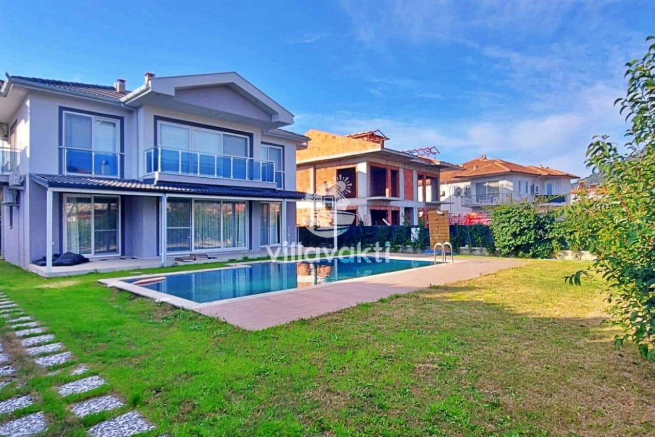 İki katlı modern villa, önünde özel havuzu ve geniş çim bahçesiyle güneşli bir günde görünüyor.