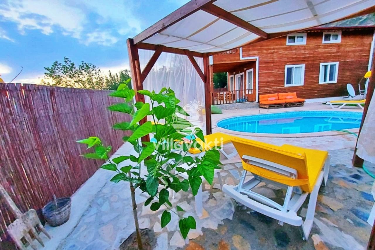 Ahşap dış cepheli villa, önünde havuz, sarı şezlonglar ve gölgelikli oturma alanı.