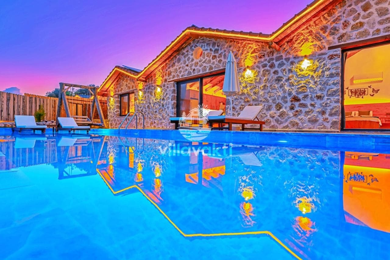 Gece ışıklandırılmış özel yüzme havuzu, taş mimarili villa dış cephesi ve şezlonglar.