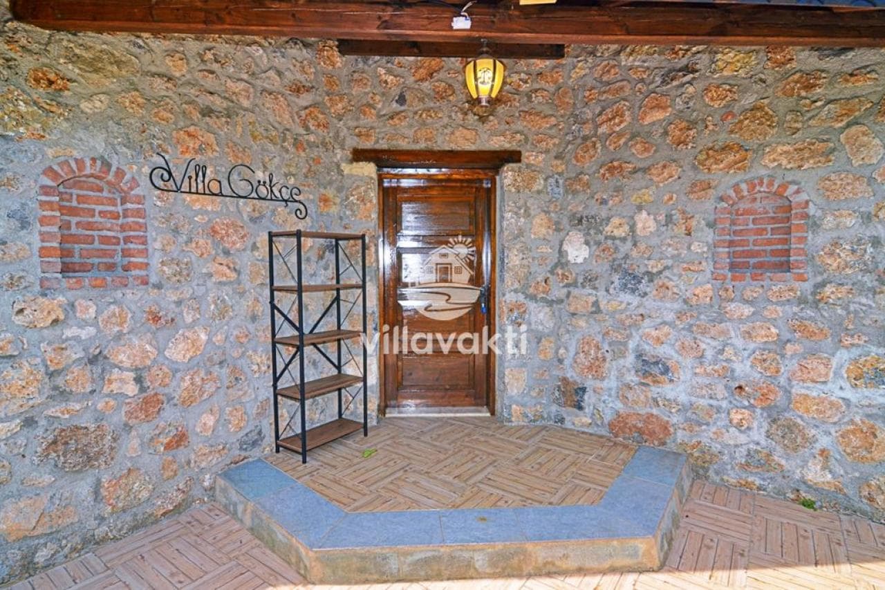 Taş duvarlı villa giriş kapısının önünde ahşap kapı, metal raf ve fener aydınlatma var.