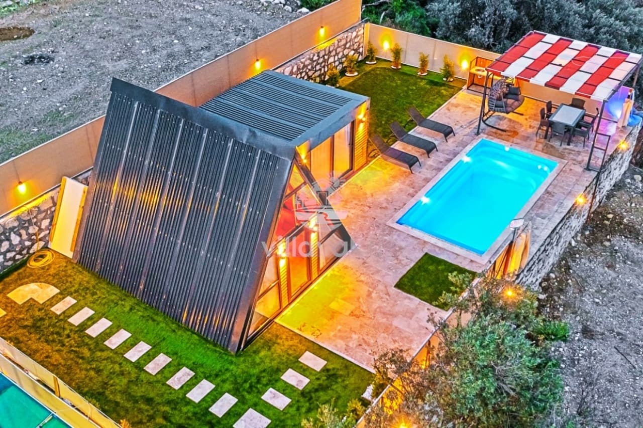 Açılı çatılı modern villanın havuzu, şezlongları ve yemek masası bulunan aydınlatmalı bahçesi.