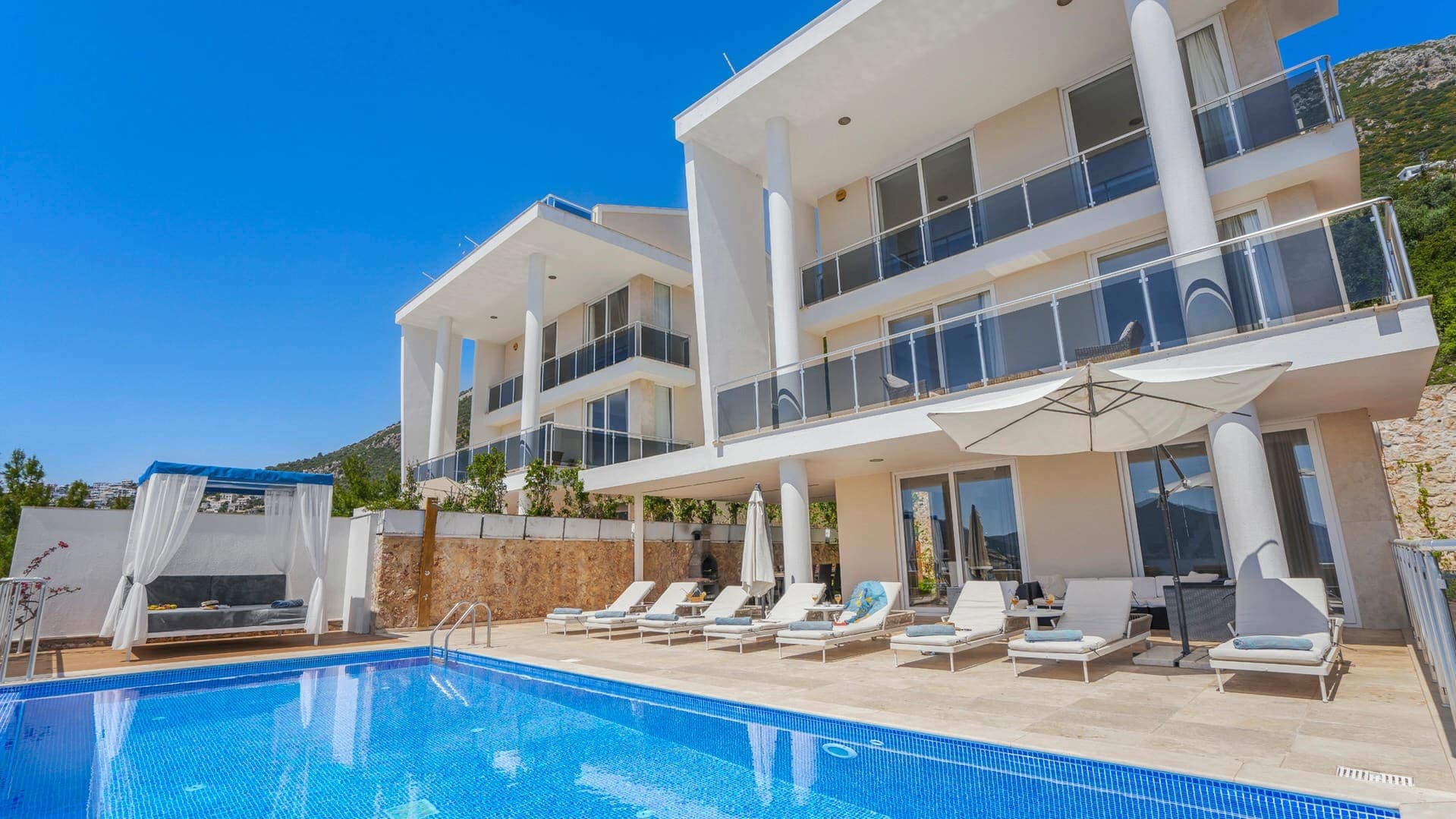 Büyük havuz kenarında şezlonglar ve şemsiyeler bulunan çok katlı modern villa.