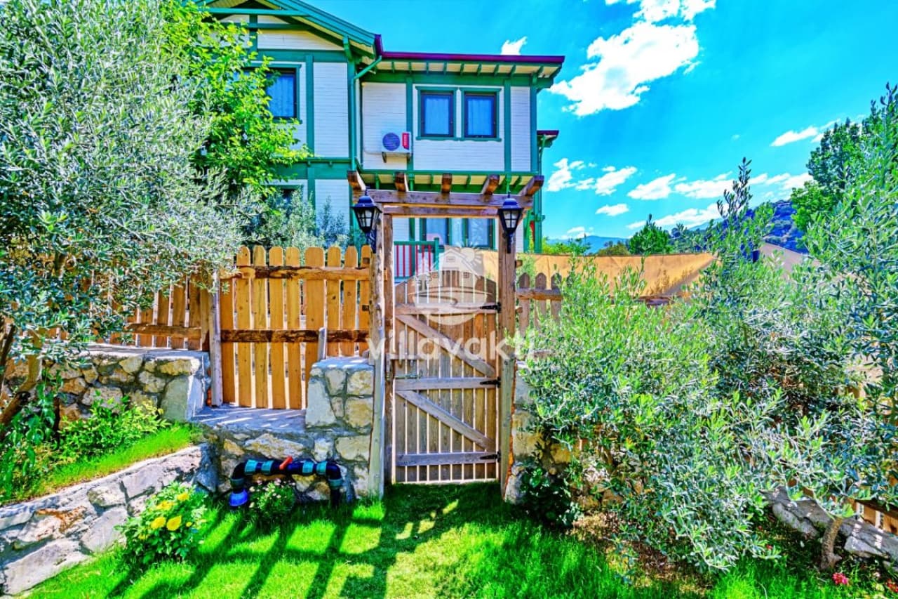 Ahşap bahçe kapısı arkasında iki katlı villa ve yeşillikler.