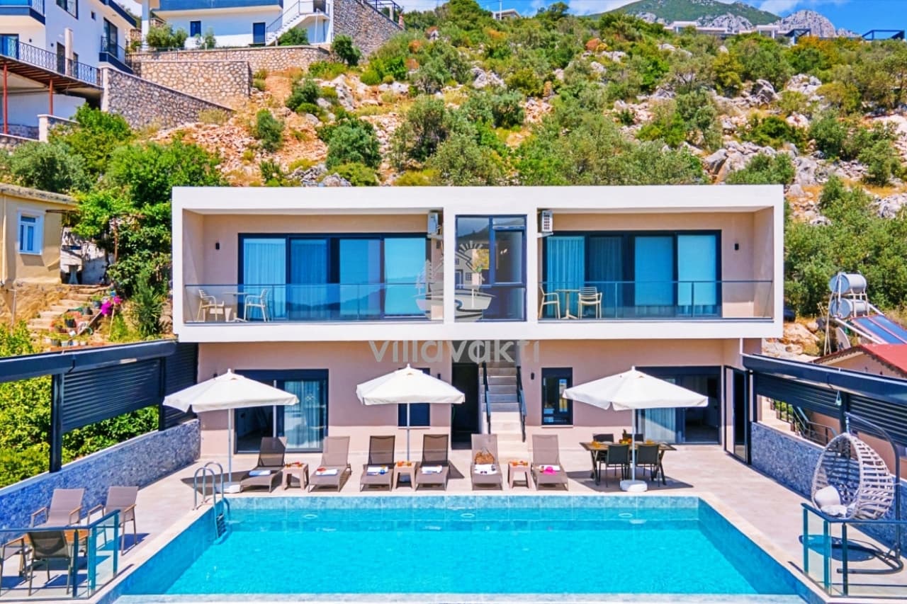 İki katlı modern villanın önünde şezlonglar, güneş şemsiyeleri ve mavi bir yüzme havuzu bulunuyor.