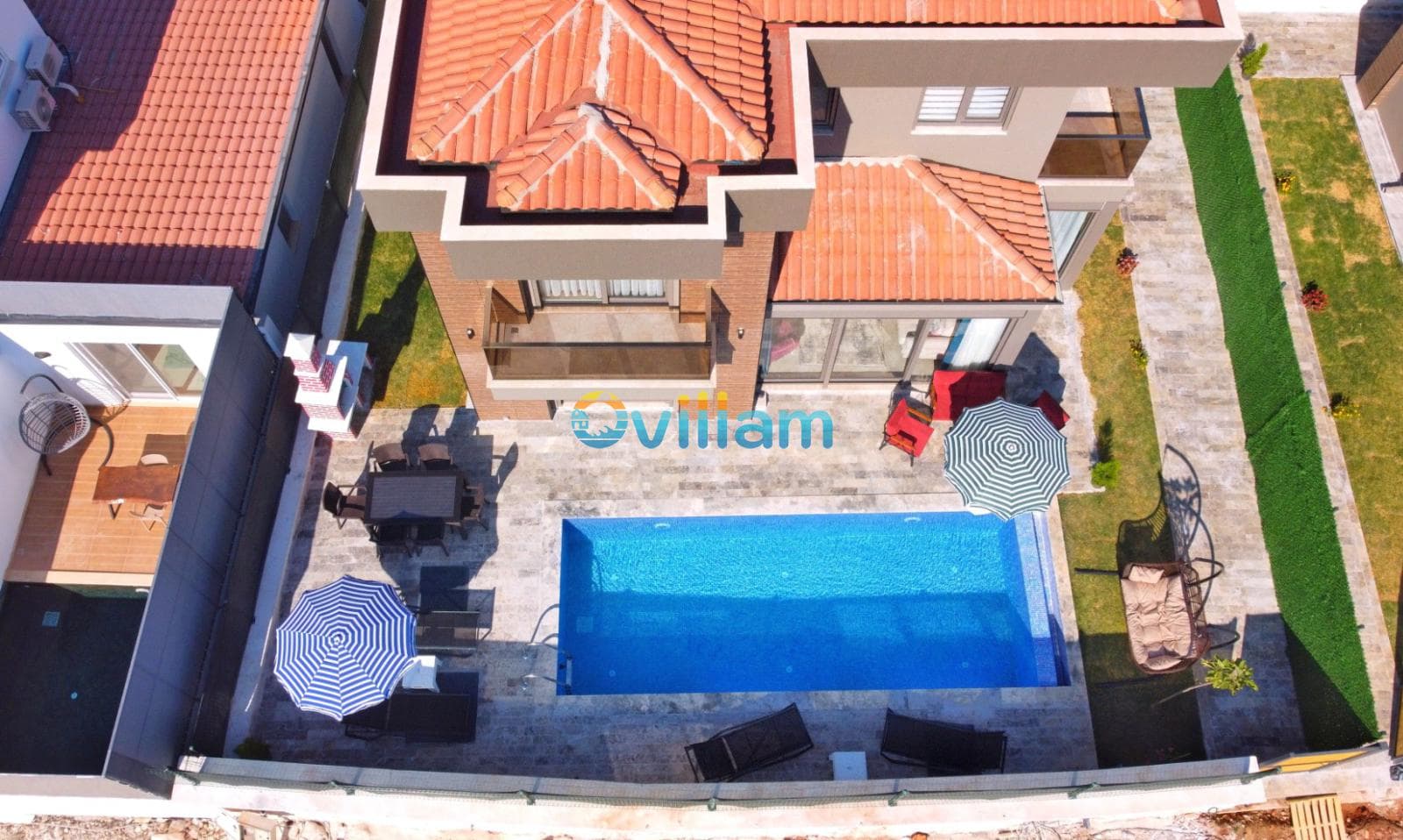 Kiremit çatılı villa, havuz, şezlonglar ve güneş şemsiyeleri ile kuş bakışı bahçe görünümü.