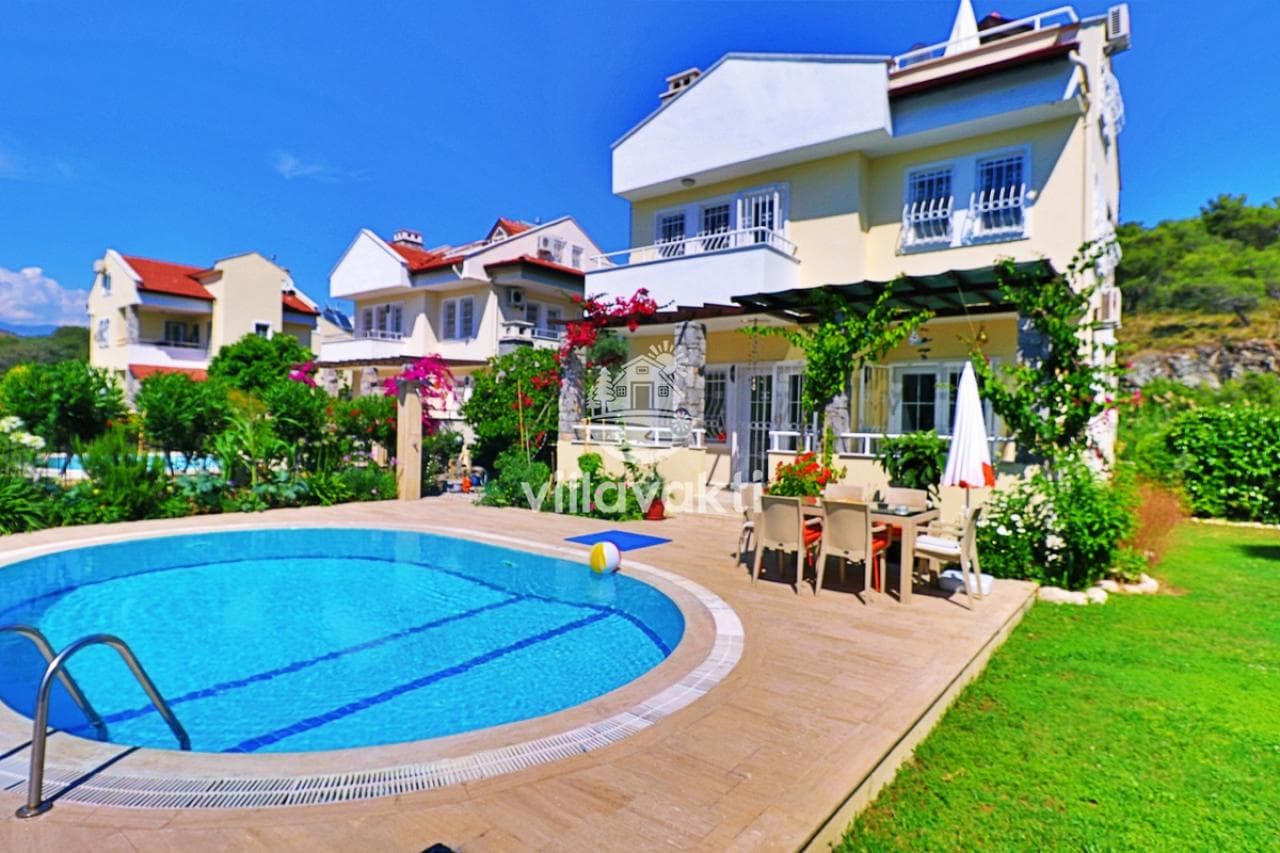 Bahçesinde yuvarlak havuz, yemek masası ve çiçekli bitkiler bulunan sarı boyalı müstakil villa.