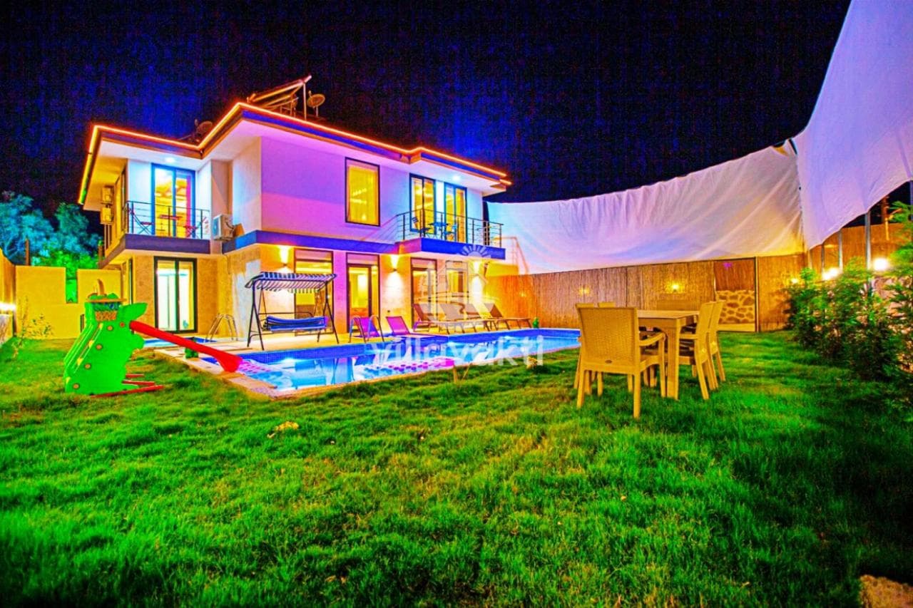 Gece aydınlatmalı, çocuk kaydıraklı havuz, bahçe oturma grubu ve villanın dış cephesi.