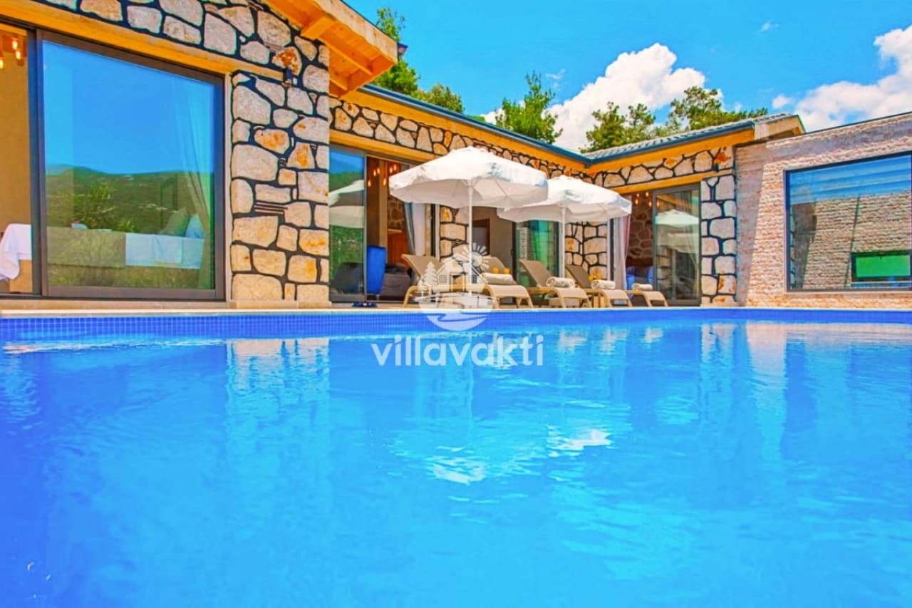 Taş duvarlı villa binası önünde şezlonglar ve güneş şemsiyeleriyle çevrili açık yüzme havuzu.