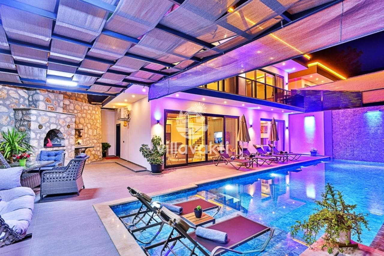Gece aydınlatmasıyla mor ve sarı ışıklar altında, havuz başında şezlonglar bulunan modern villa.