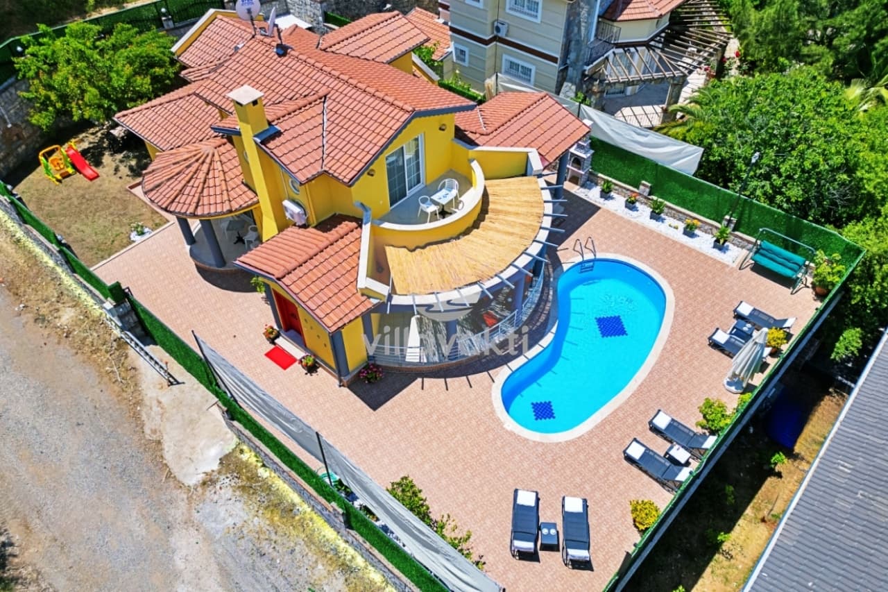 Sarı dış cepheli, kiremit çatılı villa, bahçesinde havuz ve şezlonglar.