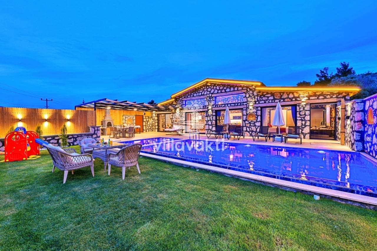 Akşam ışıklandırmalı taş duvarlı villa, önünde havuz, çim alan ve çocuk oyun grubu bulunuyor.