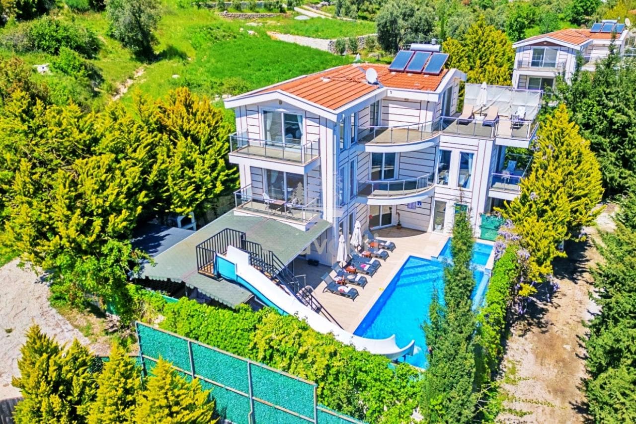 Havuz kaydırağı, güneşlenme terası ve şezlongları bulunan, yeşillikler içindeki villanın kuş bakışı görünümü.