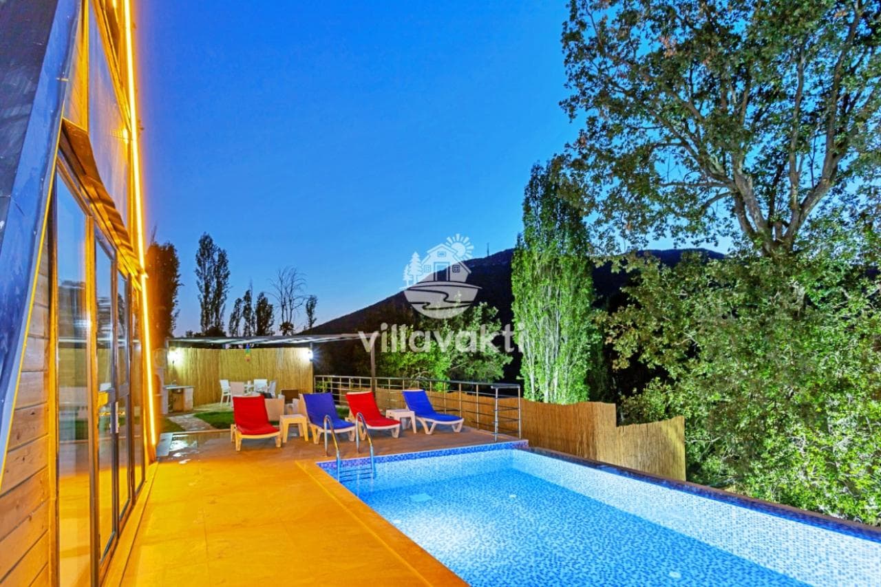 Gece aydınlatılmış A-tipi ahşap villa, önünde havuz ve şezlongların bulunduğu teras alanı.